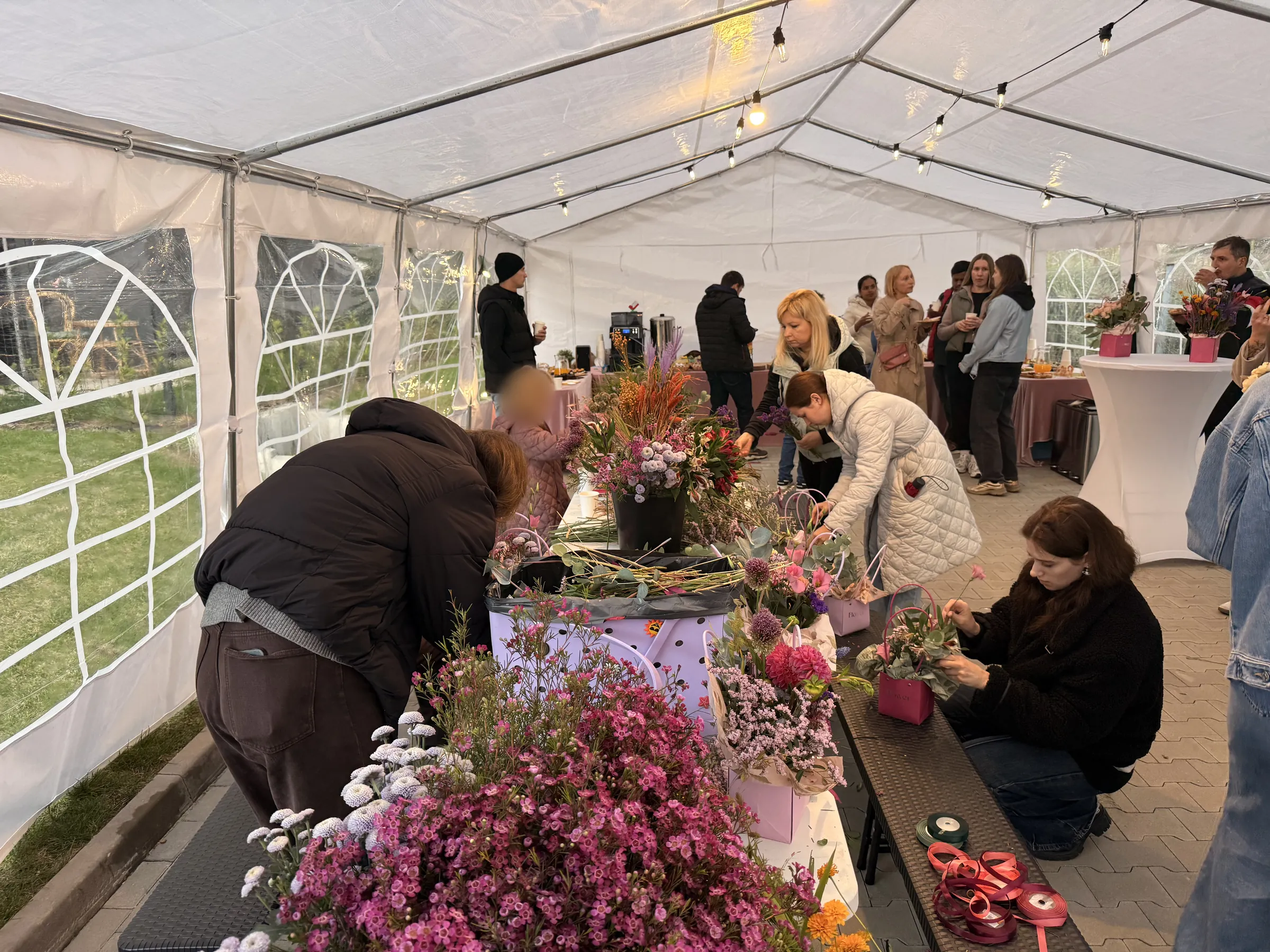 Uczestnicy układają kompozycje kwiatowe przy długim stole w namiocie ogrodowym